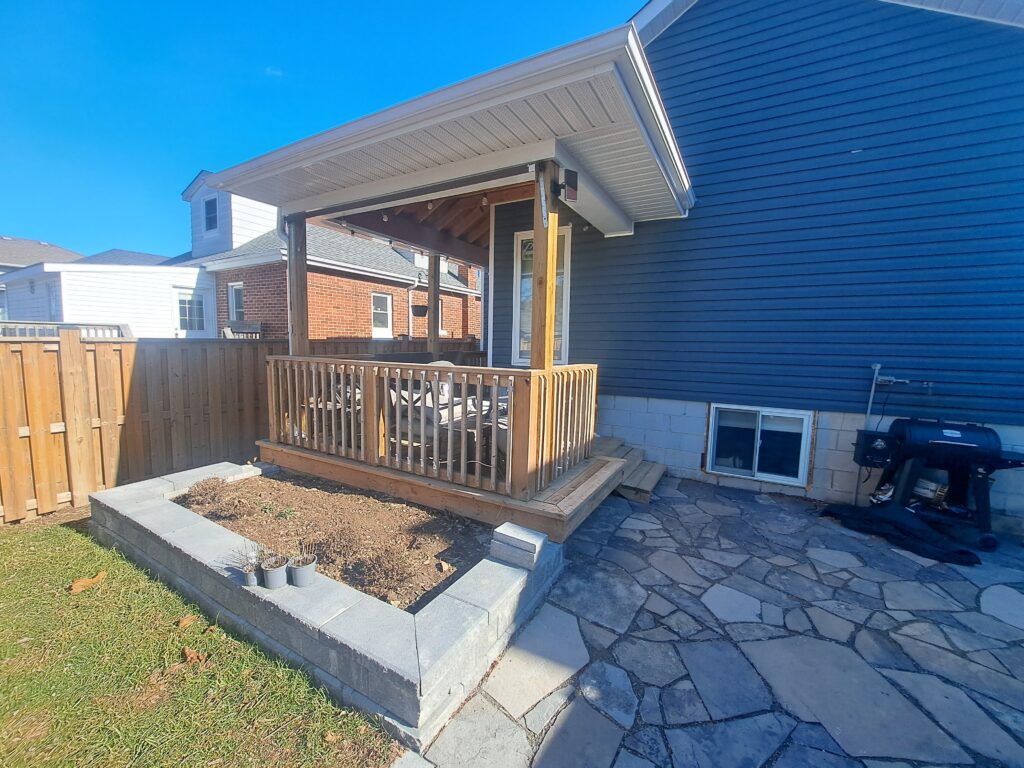 Covered backyard wood deck with railing beside a stone patio in Hamilton Ontario